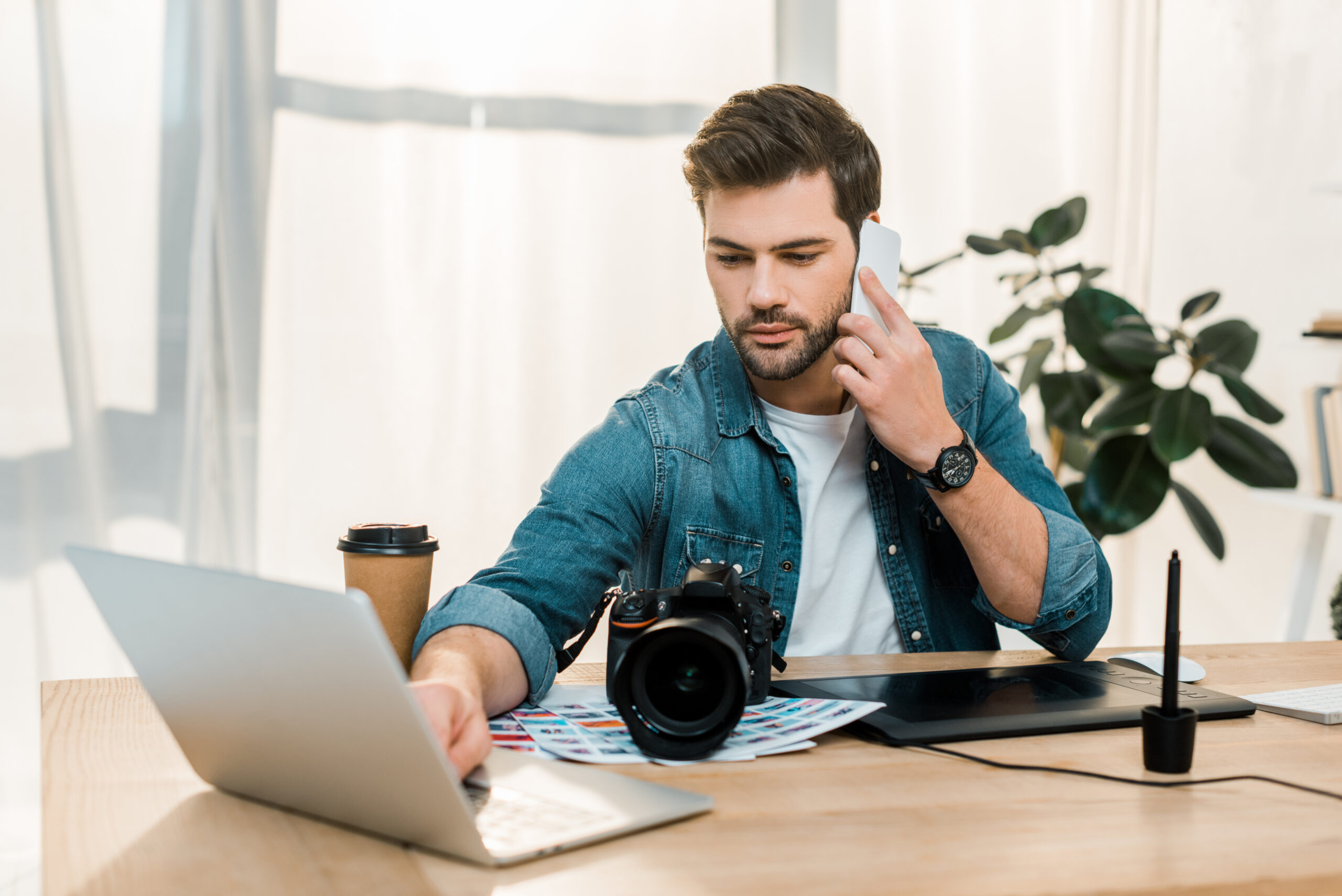 young retoucher talking by smartphone and using laptop at workplace